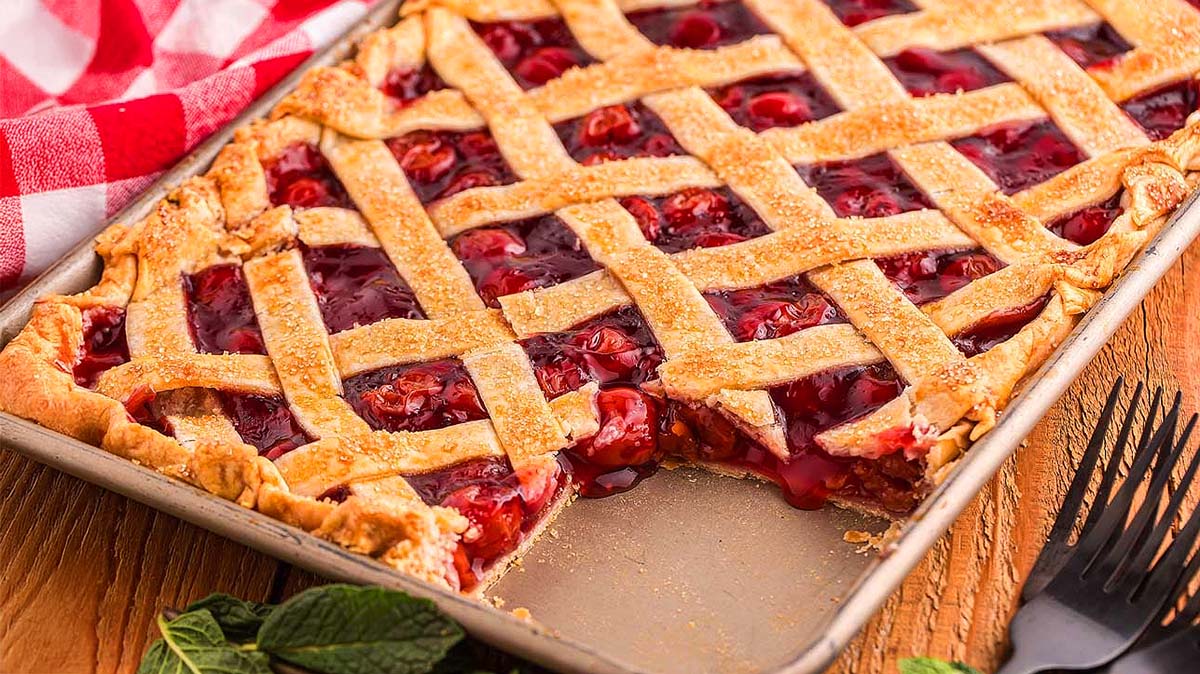 A rectangular cherry pie with a golden lattice crust in a baking pan, with one slice missing. A red and white cloth and utensils are nearby.