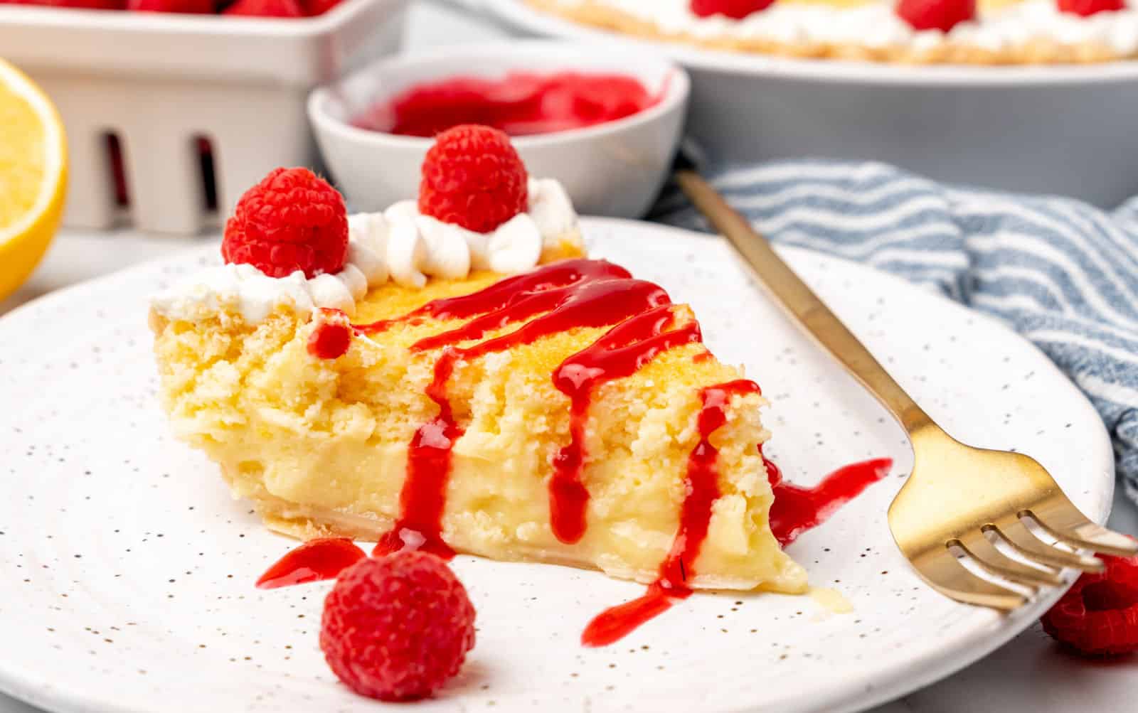 A slice of lemon pie topped with whipped cream, raspberries, and raspberry sauce sits on a white plate with a gold fork.