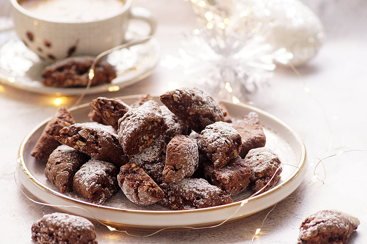 A plate of chocolate cookies dusted with powdered sugar sits on a table next to a cup of coffee and holiday decorations.