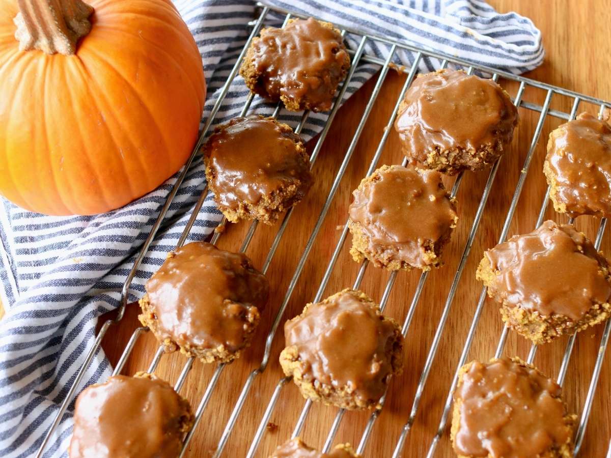 Pumpkin cookies with brown icing are cooling on a wire rack next to a whole pumpkin and a striped kitchen towel on a wooden surface.