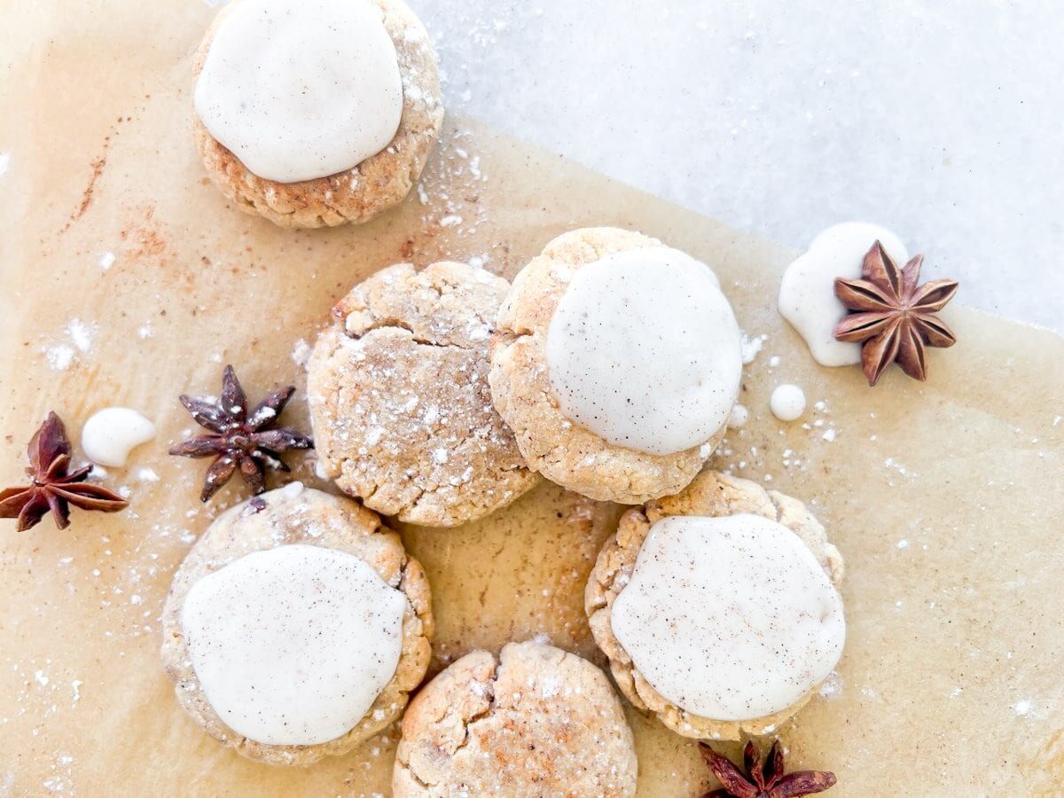 Six cookies with white icing are arranged on parchment paper, surrounded by star anise and a dusting of powdered sugar.