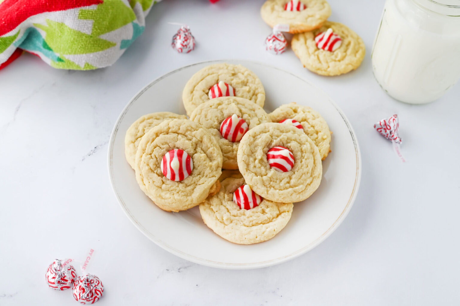 A white plate with sugar cookies topped with red and white striped peppermint candies, next to a glass of milk and a colorful towel.