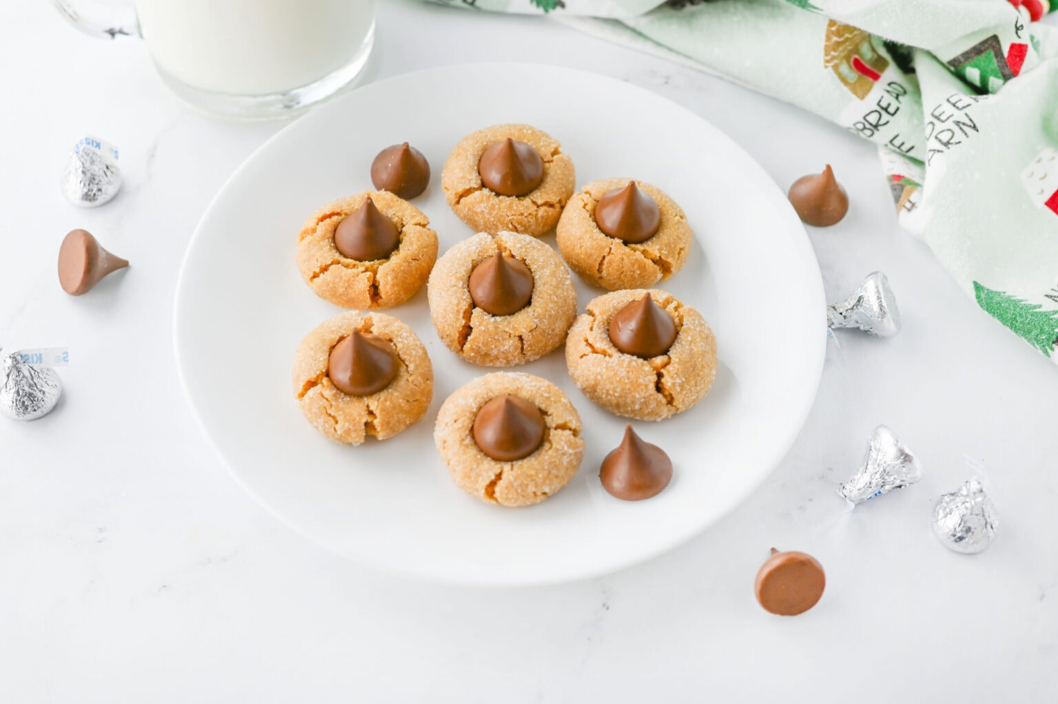 A white plate with eight peanut butter blossom cookies topped with chocolate kisses, surrounded by extra chocolate pieces, a glass of milk, and a holiday-themed napkin.