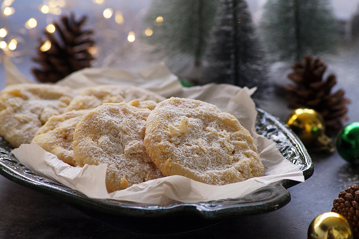 A plate lined with parchment paper holds several powdered sugar cookies, with pinecones, ornaments, and blurred festive lights in the background.