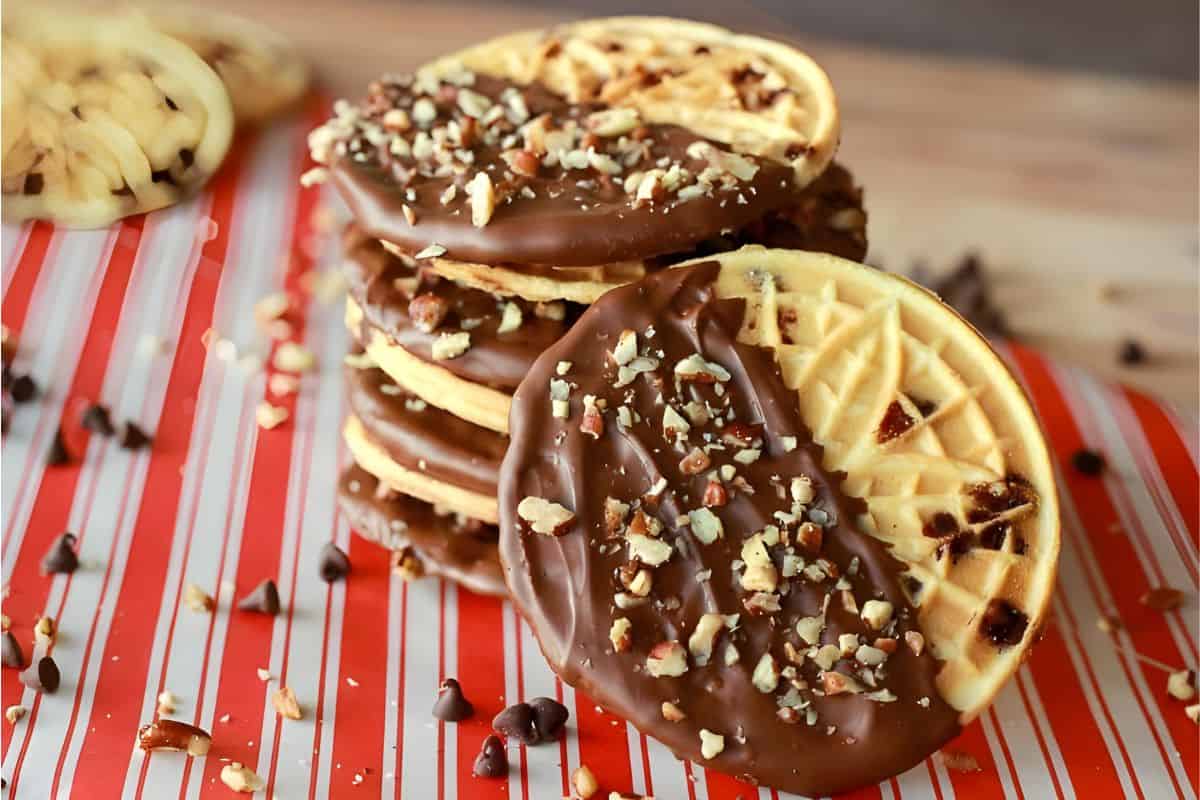 A stack of round cookies partially dipped in chocolate and sprinkled with chopped nuts, displayed on a red and white striped surface.