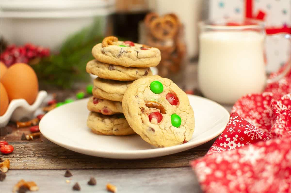 A stack of cookies with colorful candies on a white plate, next to a glass of milk, eggs, and a red festive cloth on a wooden table.