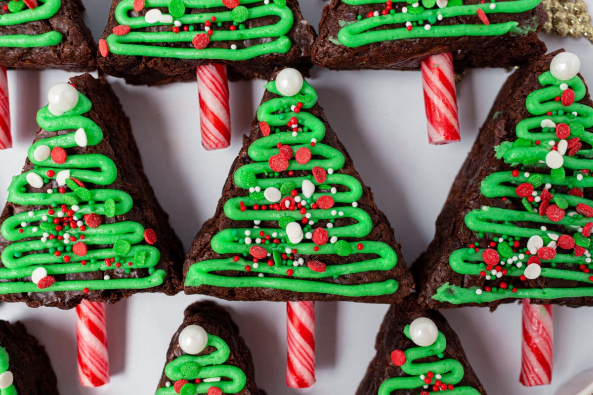 Brownies cut into triangles decorated as Christmas trees with green icing, red and white sprinkles, and candy cane sticks as trunks, arranged on a white surface.