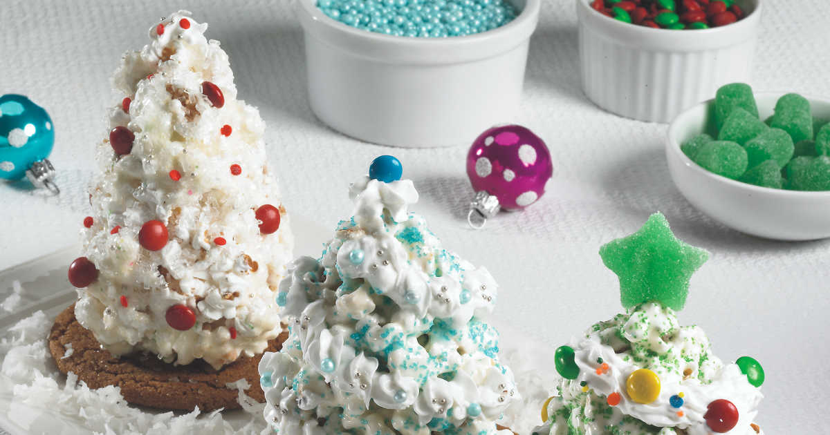 Three decorated edible Christmas trees made from cereal and icing are displayed on a white surface, with bowls of colorful candies and ornaments in the background.