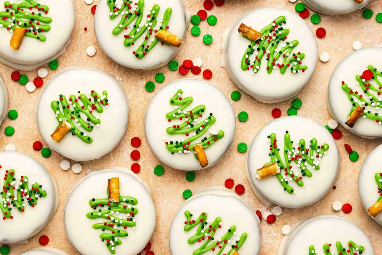 Round cookies coated in white icing, decorated with green frosting Christmas trees, multicolored sprinkles, and pretzel sticks as trunks, arranged on a beige surface.