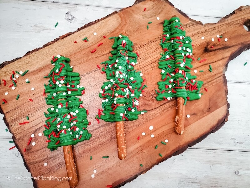 Three pretzel sticks decorated as Christmas trees with green icing and red and white sprinkles are arranged on a wooden cutting board.