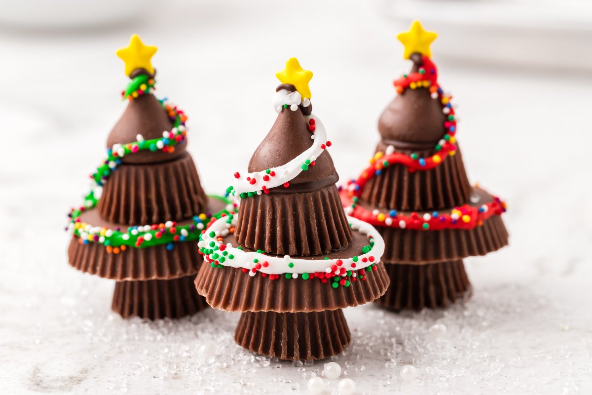 Three Christmas tree-shaped treats made from stacked chocolate peanut butter cups, topped with chocolate kisses and decorated with sprinkles, icing, and yellow star candies.
