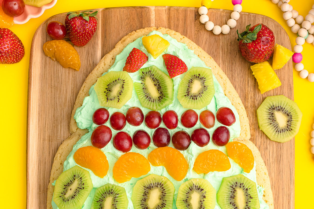 A large cookie in the shape of an Easter egg topped with green frosting, sliced kiwis, strawberries, grapes, orange segments, and pineapple pieces on a wooden board.