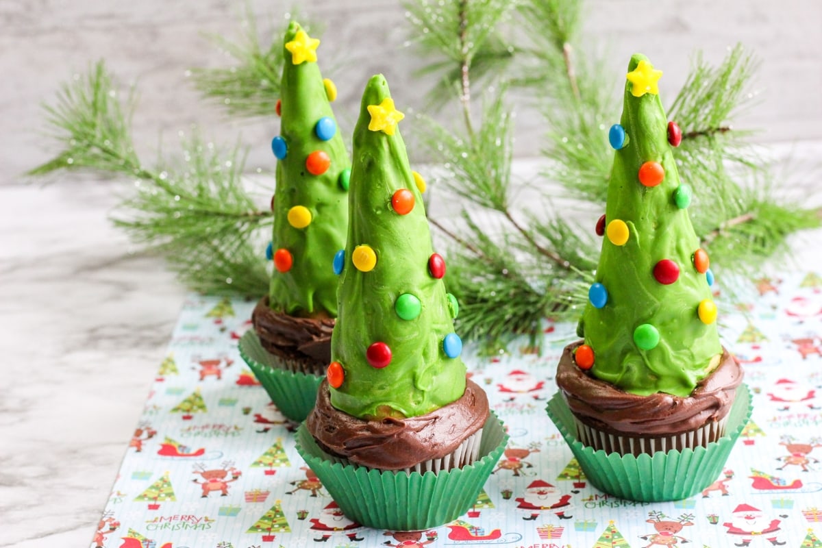 Three cupcakes topped with green frosting shaped like Christmas trees, decorated with colorful candy dots and yellow stars, placed on a festive holiday-themed surface.