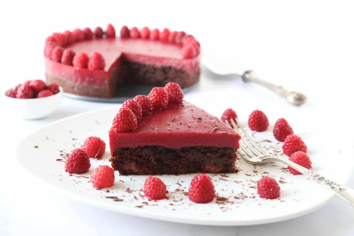 A slice of chocolate and raspberry cake is served on a plate with fresh raspberries and chocolate shavings, with the remaining cake and a cake server in the background.