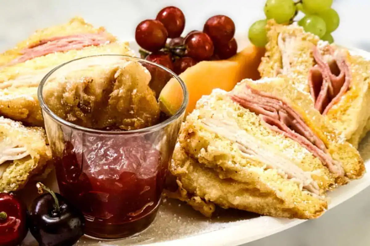 Plate with Monte Cristo sandwich halves, red and green grapes, cantaloupe slices, cherries, and a small glass of red jam for dipping.