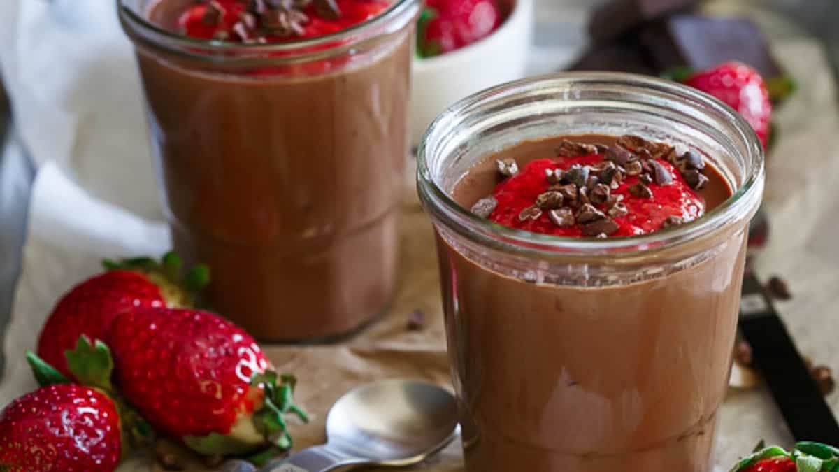 Two glass jars filled with chocolate mousse, topped with strawberry sauce and cacao nibs, surrounded by fresh strawberries and chocolate pieces.