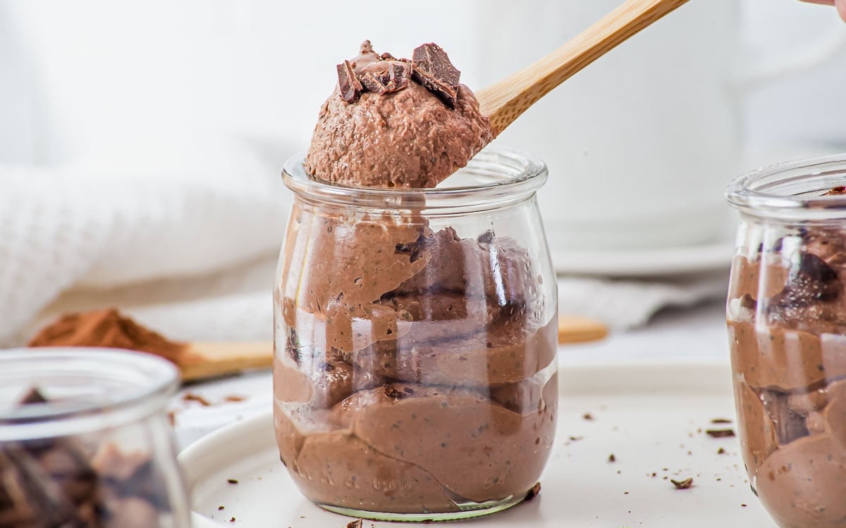 A wooden spoon holds up a scoop of chocolate mousse from a glass jar, with chopped chocolate pieces on top.