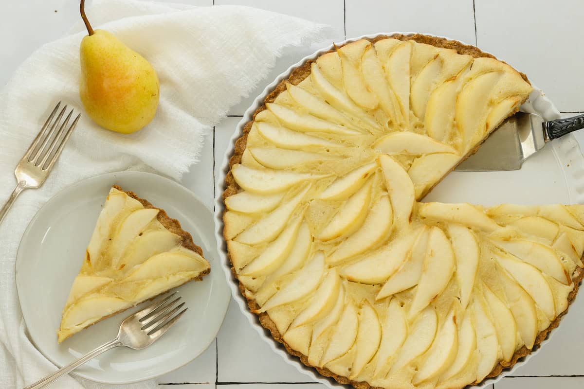 A pear tart with several slices missing sits on a white tiled surface next to a plate holding a single slice, two forks, a white napkin, and a fresh pear.