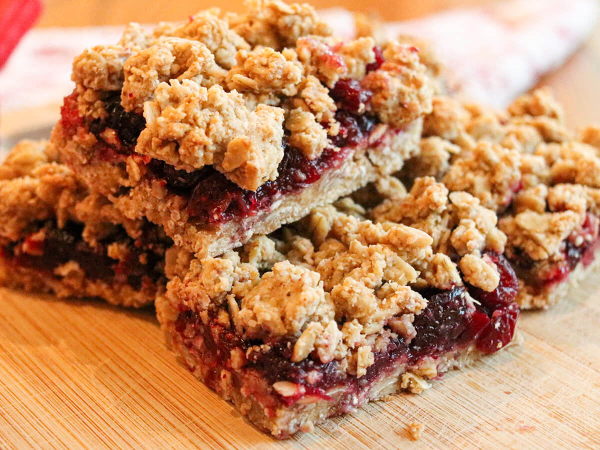 Three oat and fruit crumble bars stacked on a wooden surface, showing layers of crumbly oats and a red fruit filling.