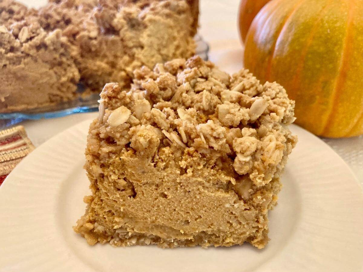 A slice of pumpkin cheesecake with an oat crumble topping is served on a white plate, with a whole cheesecake and pumpkins in the background.