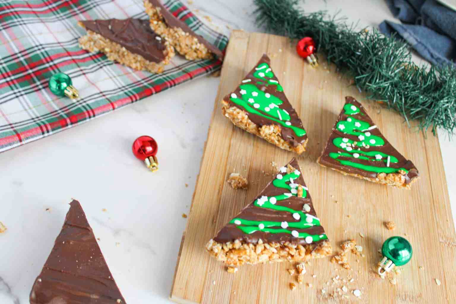 Triangular chocolate-topped treats with green icing and sprinkles are arranged on a wooden board, surrounded by small ornaments and a plaid cloth.