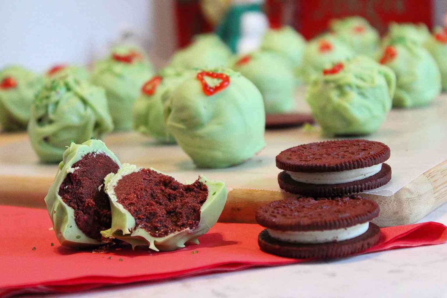 Red velvet cake truffles coated in green chocolate sit beside chocolate sandwich cookies on a red napkin, with more truffles in the background.