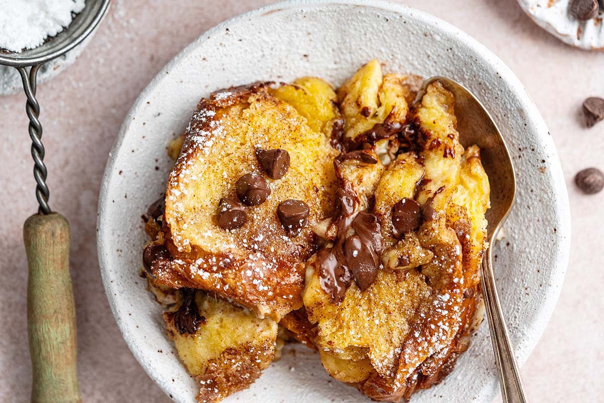 A plate of French toast topped with chocolate chips and powdered sugar, served with a spoon on a white plate.
