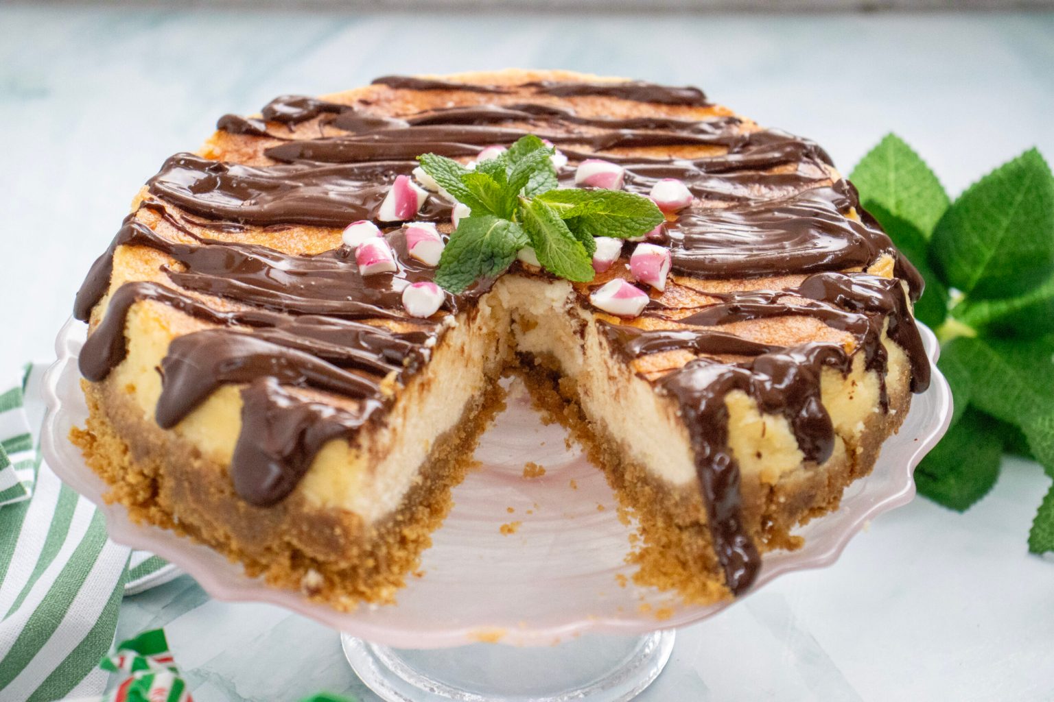A cheesecake with a graham cracker crust, topped with chocolate drizzle, crushed peppermint candies, and a fresh mint garnish, with one slice removed.