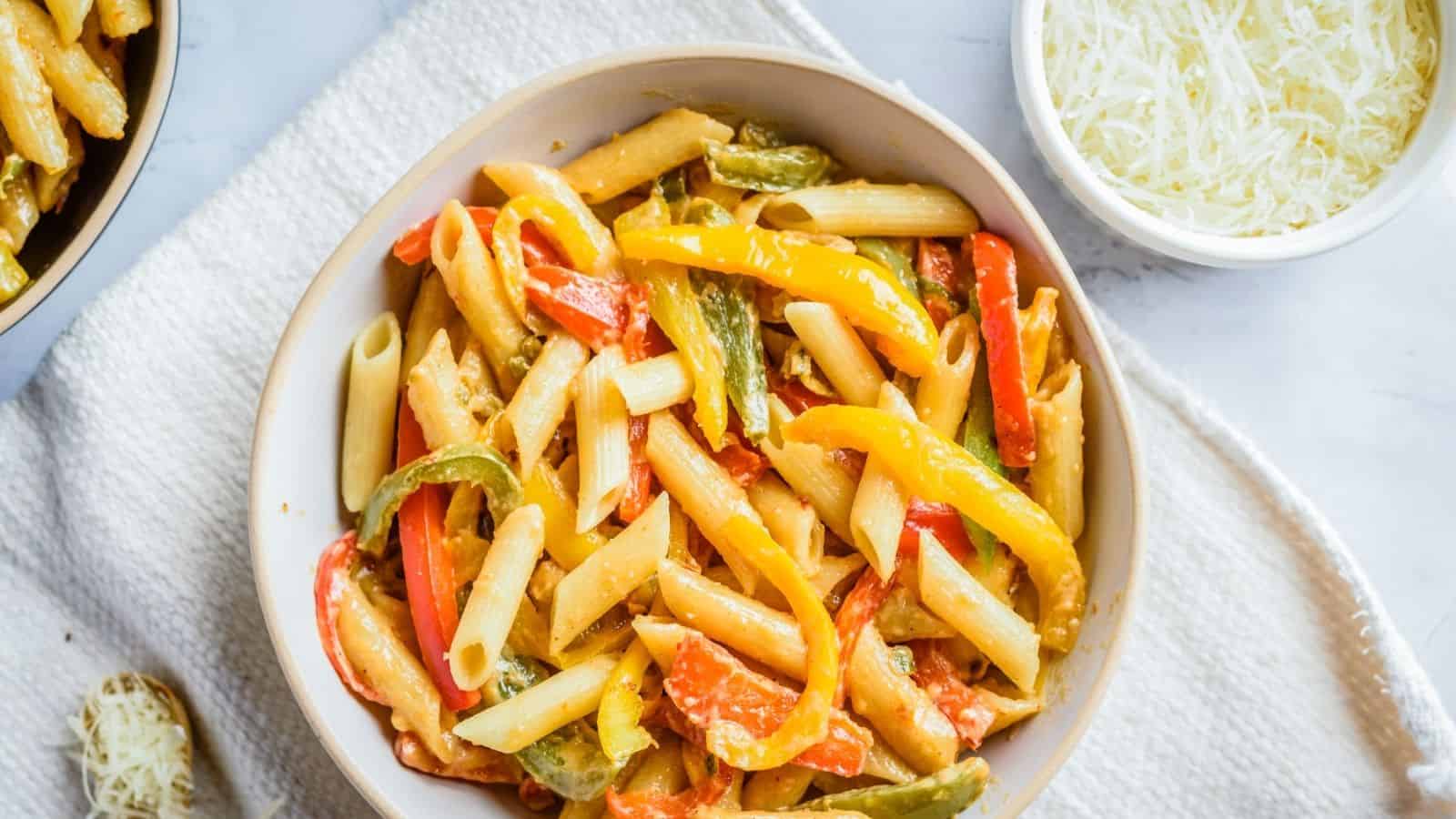 Bowl of penne pasta mixed with sliced red, yellow, and green bell peppers, next to a bowl of grated cheese on a light surface.