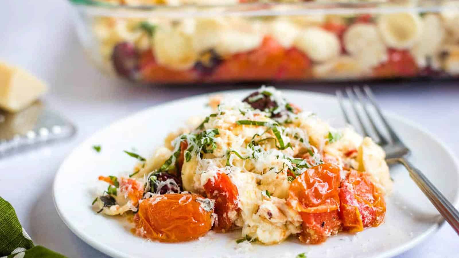 A serving of baked pasta with cherry tomatoes, feta cheese, and herbs on a white plate with a fork; a casserole dish is visible in the background.