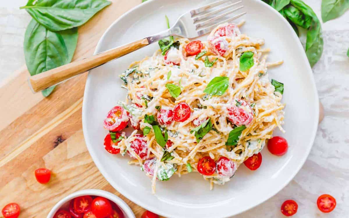 A plate of pasta mixed with cherry tomatoes, fresh basil, and a creamy white sauce, served with a fork on a white plate.