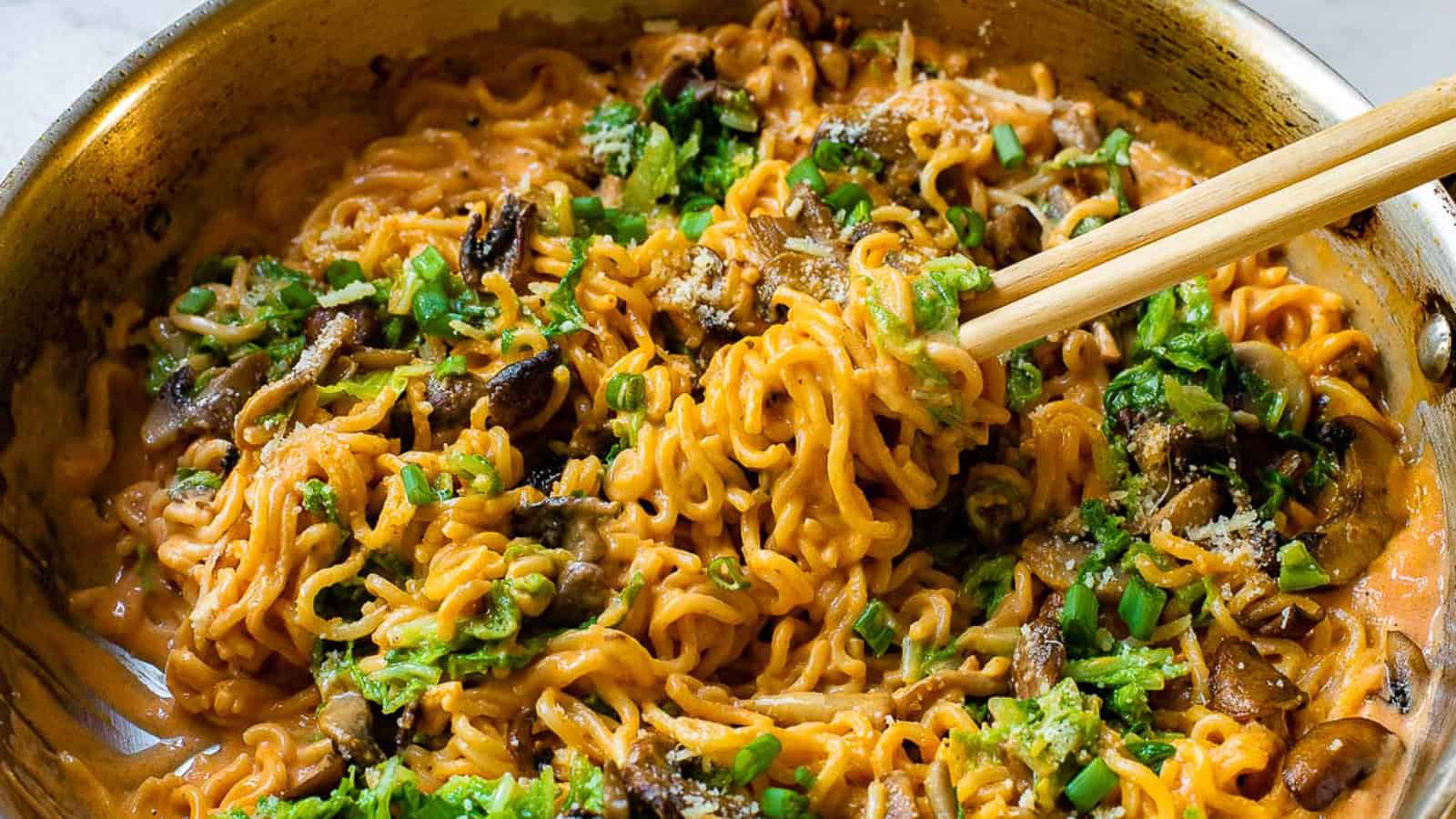 A pan of cooked ramen noodles with mushrooms, green herbs, and sauce, being lifted with chopsticks.