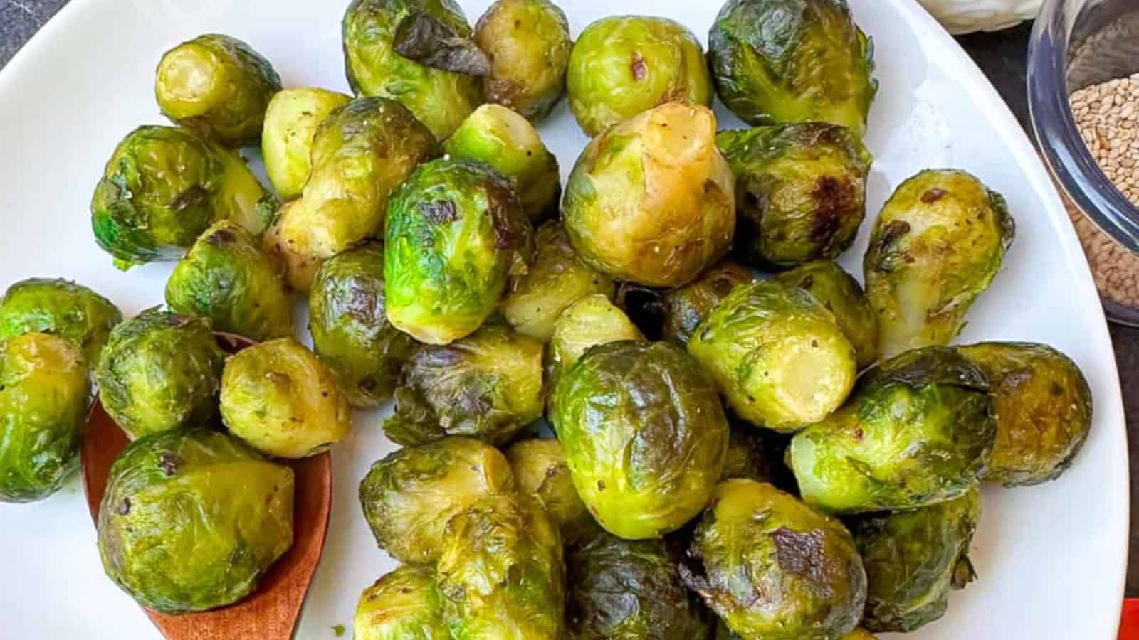 A plate of cooked Brussels sprouts with a wooden spoon underneath some of the vegetables, next to a glass bowl containing sesame seeds.