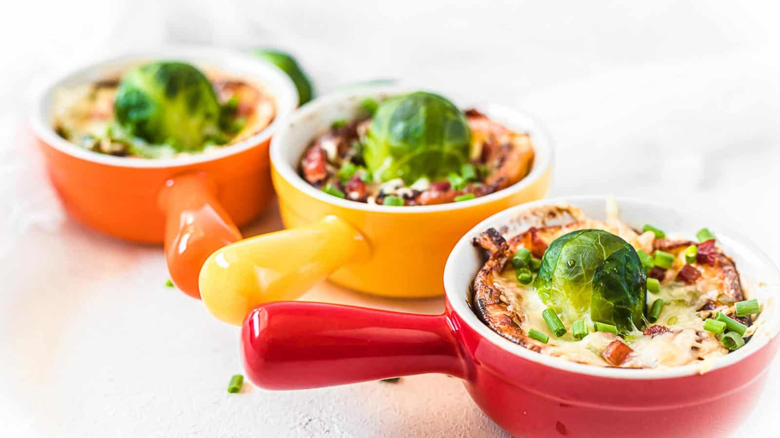 Three small colorful baking dishes filled with a baked casserole, each topped with a Brussels sprout and garnished with chopped chives.