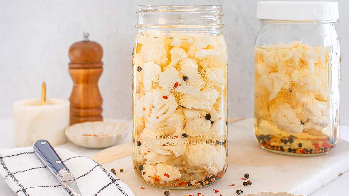 Two glass jars filled with pickled cauliflower and spices sit on a white surface, with a pepper grinder, dish, candle, and kitchen towel nearby.