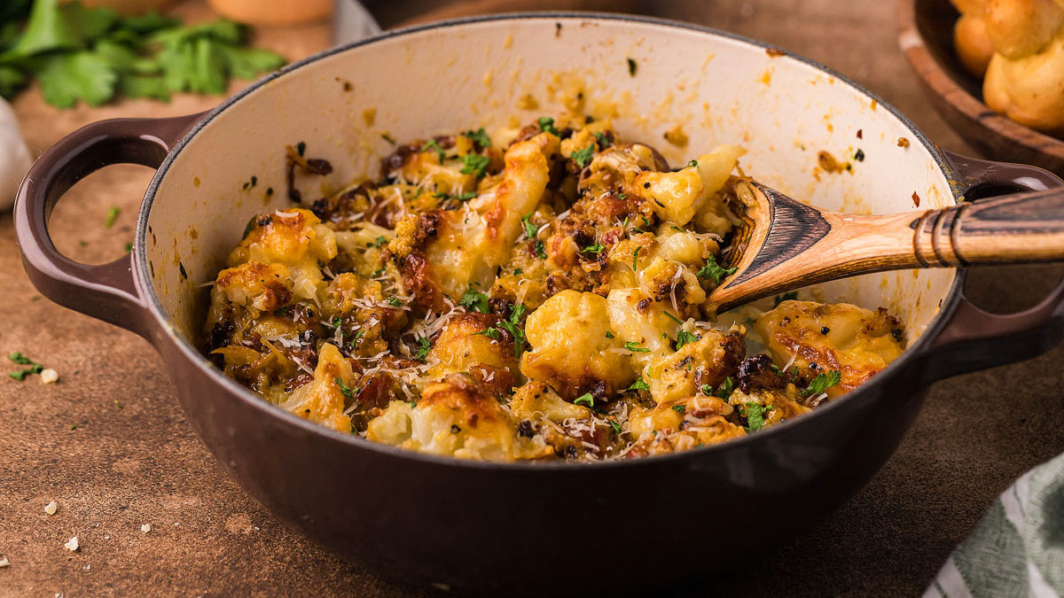 A brown casserole dish filled with baked cauliflower, cheese, herbs, and a wooden spoon resting inside.