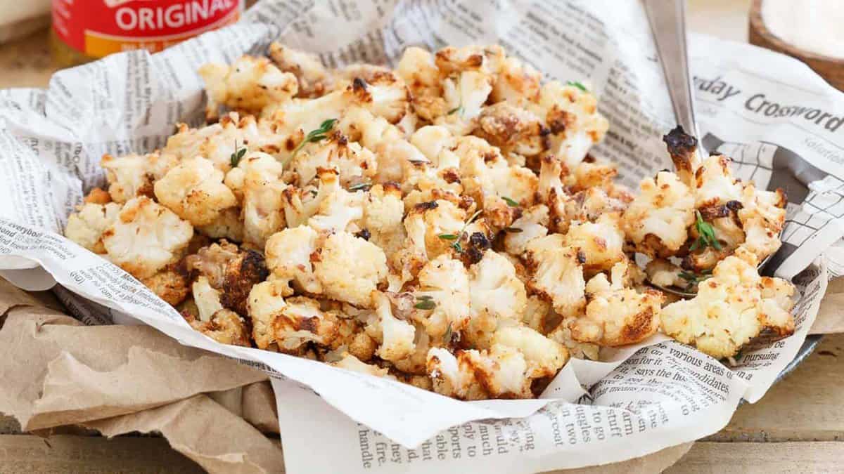 A basket lined with newspaper holds roasted cauliflower pieces garnished with herbs, with a fork resting on the side.