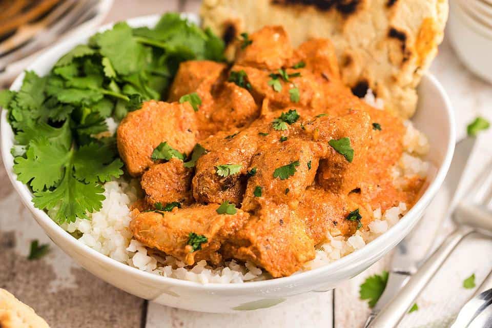 A bowl of chicken tikka masala served over rice, garnished with fresh cilantro and accompanied by a piece of naan bread.