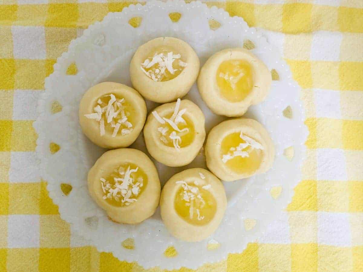 A white plate with seven lemon thumbprint cookies topped with shredded coconut, placed on a yellow and white checkered tablecloth.