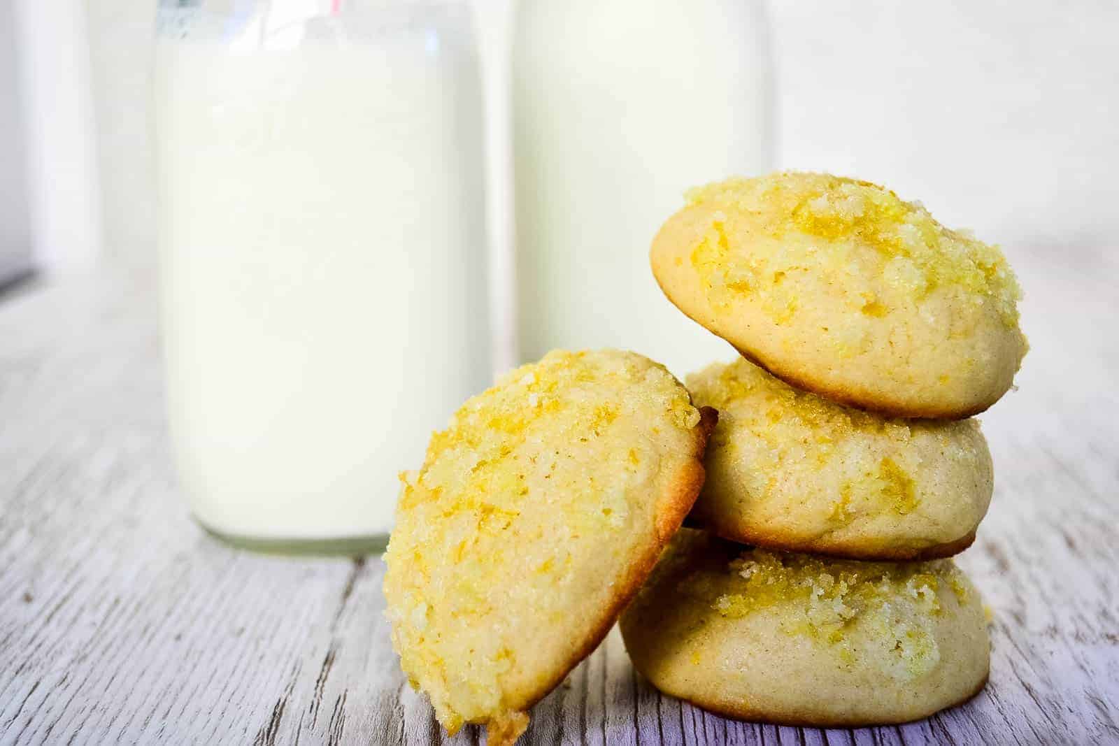 Four lemon sugar cookies are stacked next to two glass bottles of milk on a light wooden surface.