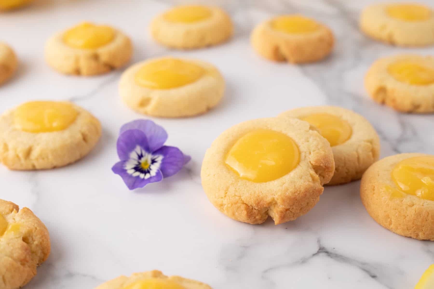 Round cookies with yellow filling are arranged on a marble surface, with a purple flower placed among them.