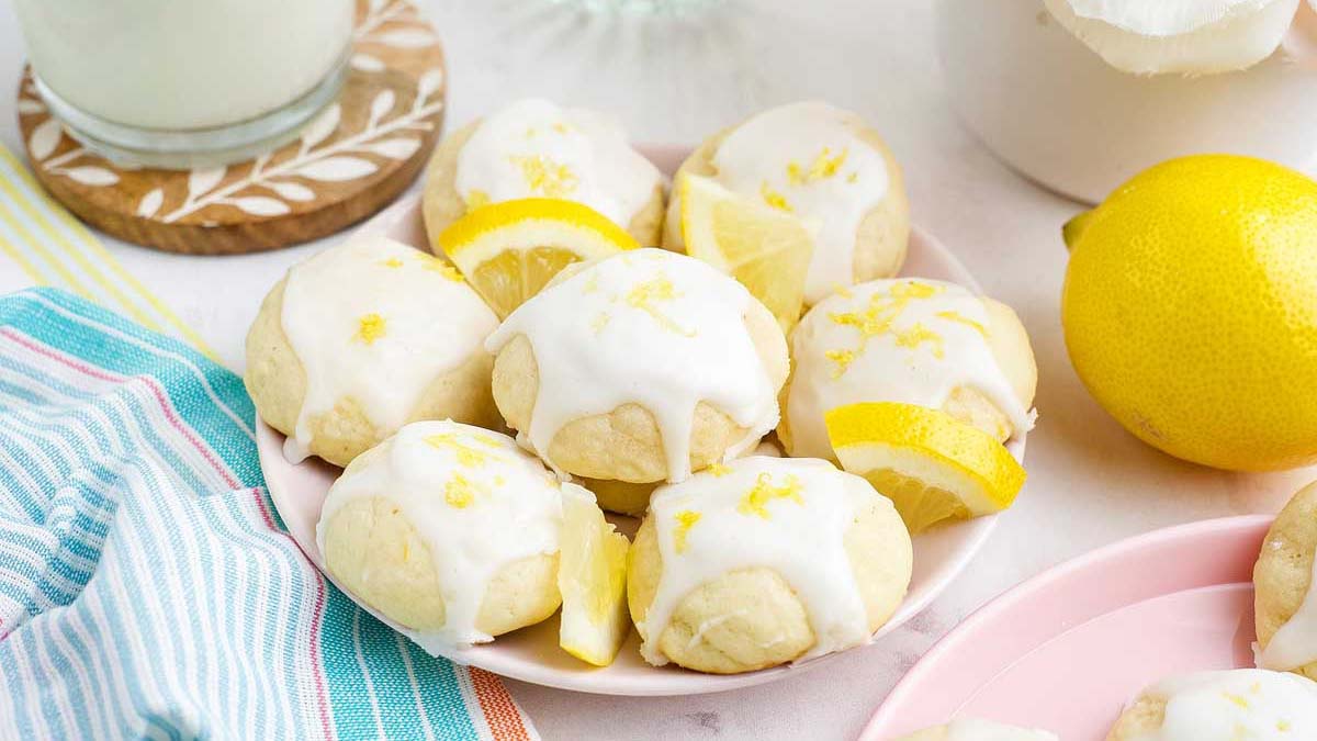 A plate of lemon cookies topped with white icing and lemon zest, garnished with lemon wedges, next to a glass of milk and a whole lemon.