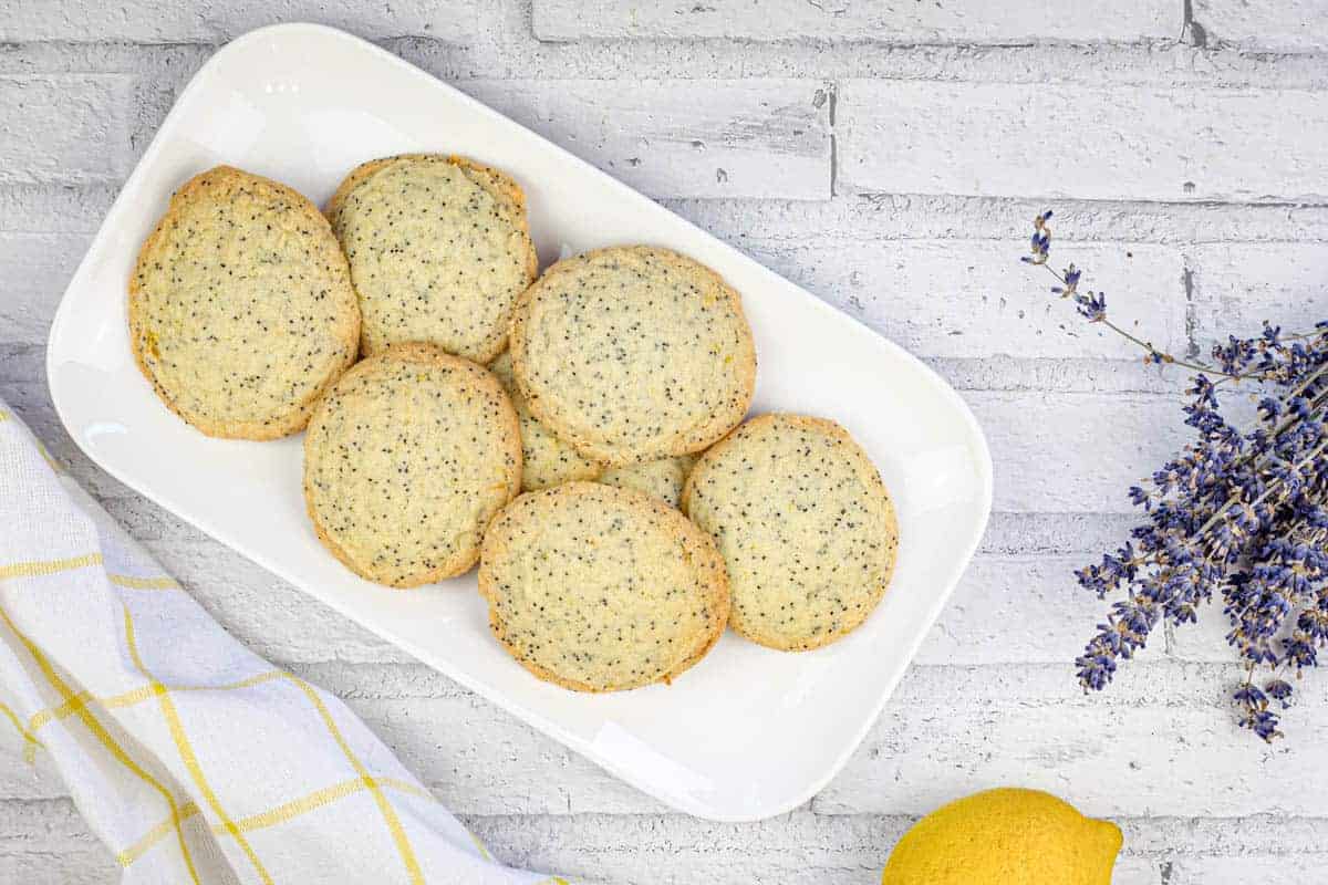 A white rectangular plate holds seven round poppy seed cookies, placed on a light brick surface with a lemon, a lavender sprig, and a checkered towel nearby.