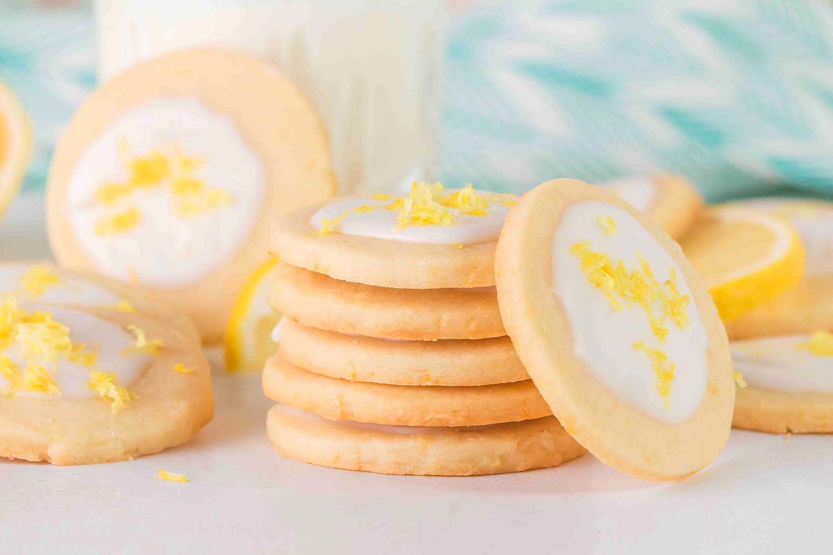 A stack of lemon cookies with white icing and lemon zest on top, surrounded by more cookies and lemon slices.