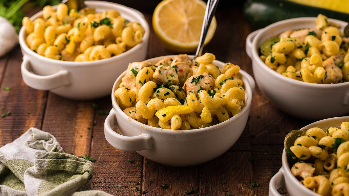 Three white bowls filled with pasta, chicken, and parsley on a wooden table, with a lemon half and a green cloth nearby.