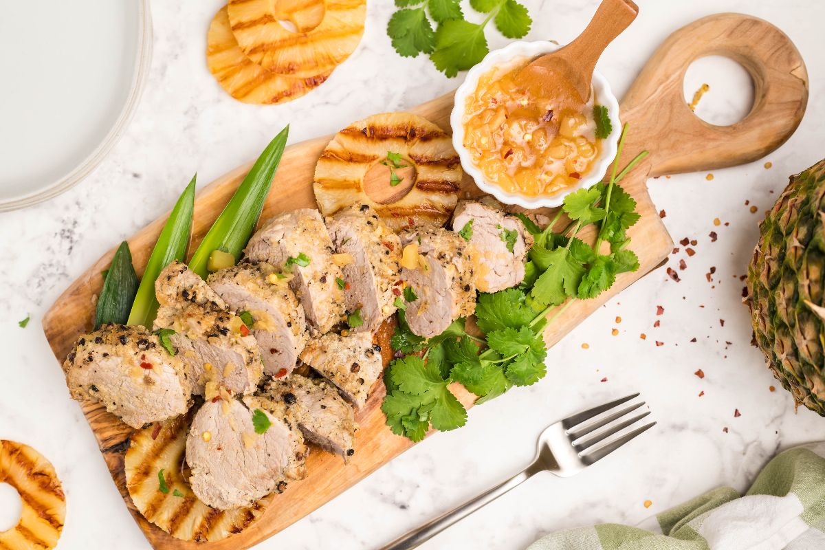 Sliced seasoned pork with grilled pineapple, fresh cilantro, okra, and a bowl of sauce on a wooden board, placed on a marble surface with a fork nearby.