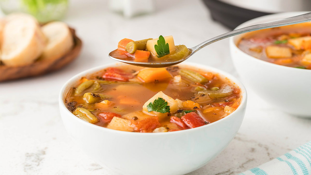 A bowl of vegetable soup with diced carrots, celery, potatoes, and tomatoes, topped with parsley, with a spoonful of soup held above the bowl. Sliced bread is in the background.