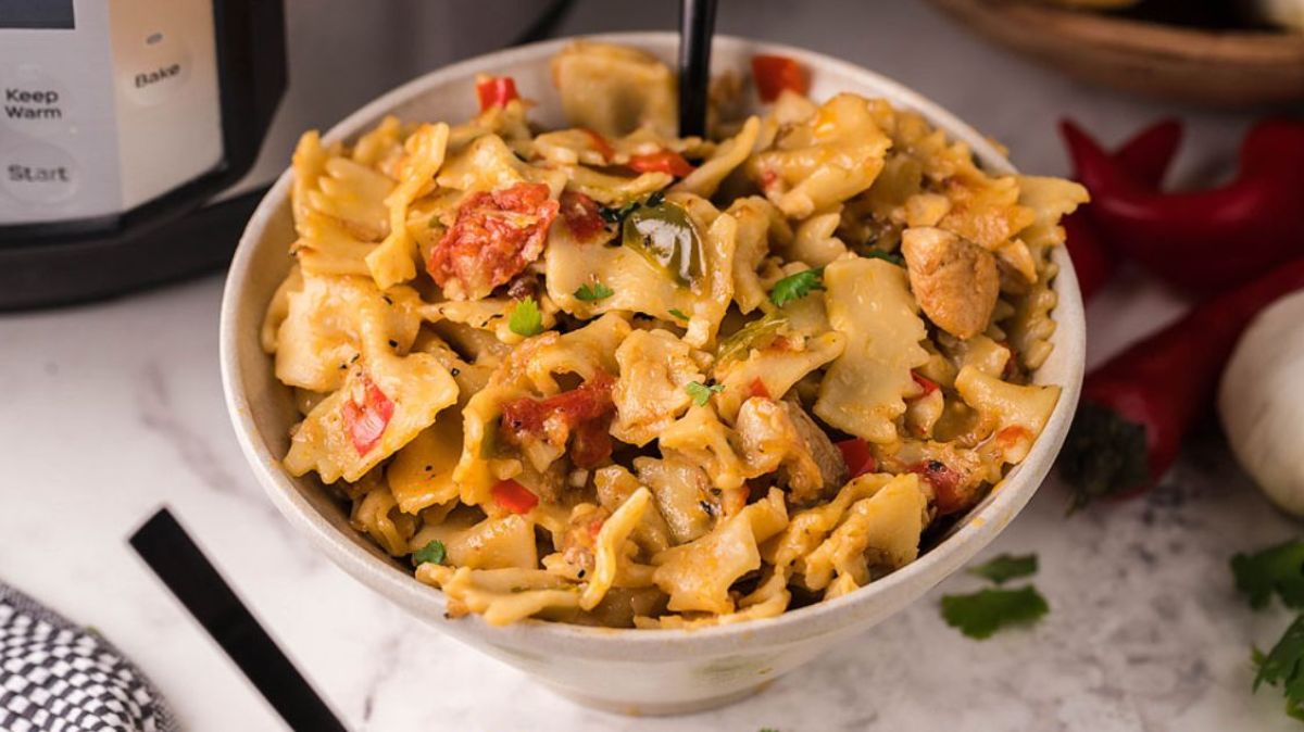 A bowl of bow-tie pasta mixed with pieces of chicken, red peppers, and a creamy sauce, garnished with chopped herbs.