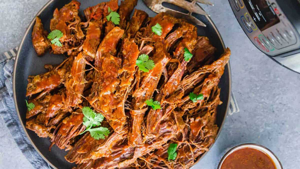 A plate of shredded beef barbacoa garnished with cilantro, with a fork, a slow cooker, and a bowl of sauce nearby on a gray surface.