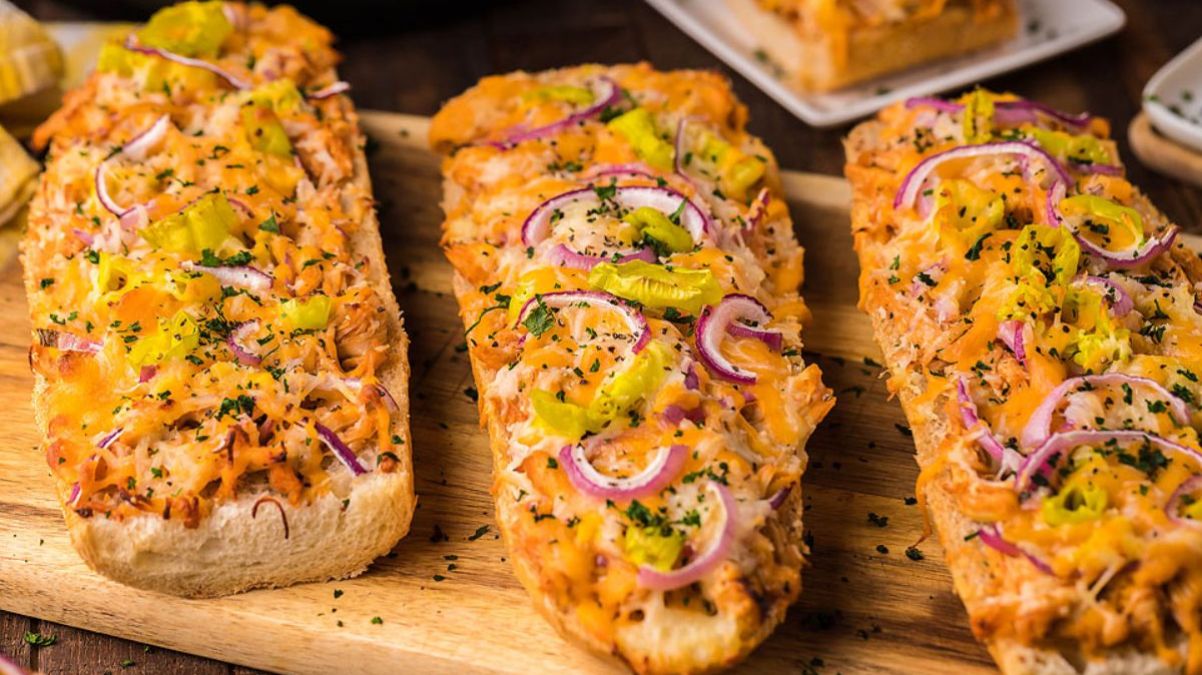 Three pieces of cheesy bread topped with shredded chicken, sliced red onions, banana peppers, and chopped herbs on a wooden cutting board.